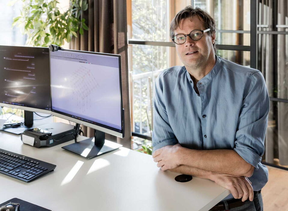 Ein Mann mit Brille sitzt an einem Schreibtisch mit zwei Monitoren, die technische Pläne anzeigen.