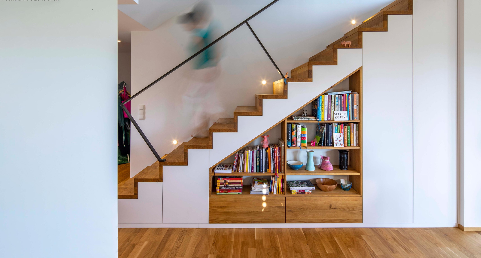 Eine Person läuft eine elegante Holztreppe hinauf, während ein Bücherregal darunter sichtbar ist.
