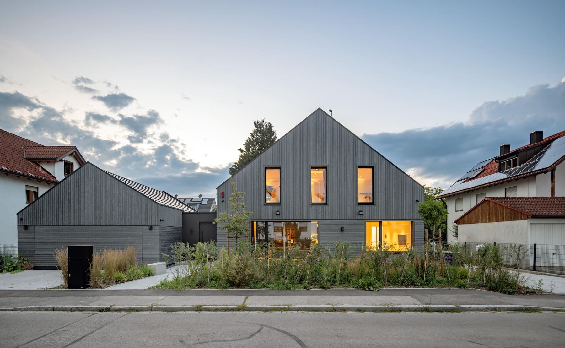 Ein modernes, graues Haus mit großen Fenstern und grüner Bepflanzung strahlt im Abendlicht.