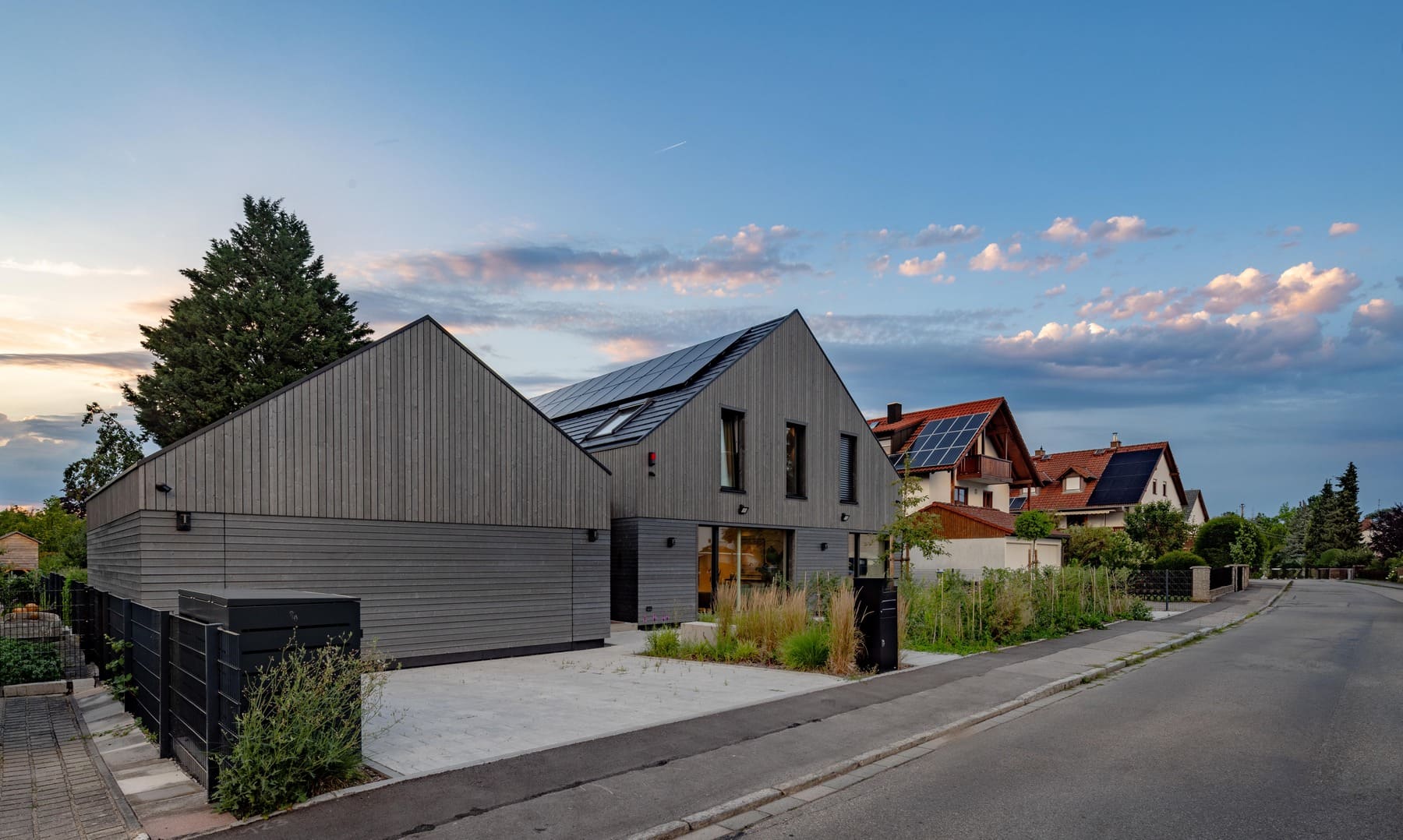 Modernes Haus mit Holzfassade und Solarpanelen, umgeben von grüner Landschaft und angrenzenden Straßen.