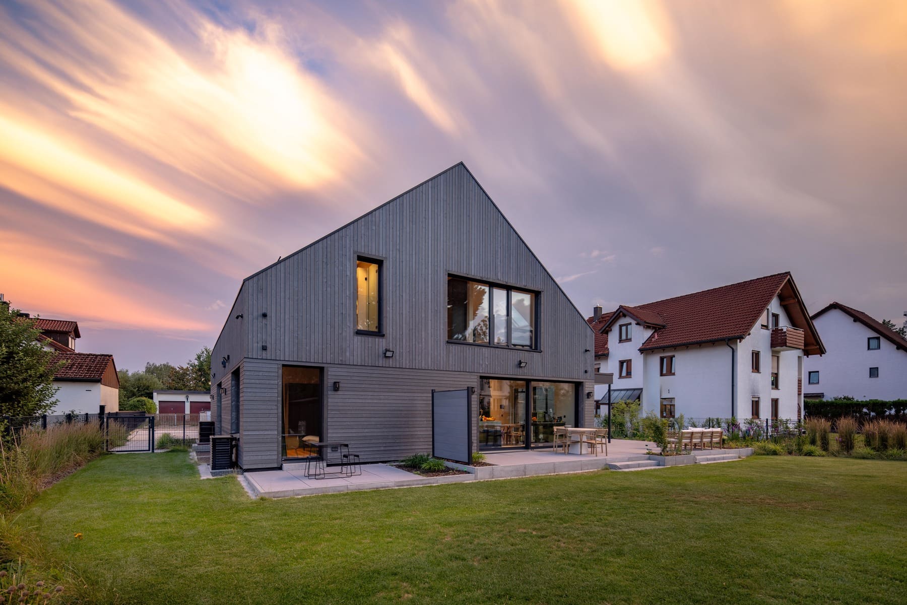 Modernes Haus mit Holzverkleidung und Garten, unter einem dramatischen Himmel bei Sonnenuntergang.