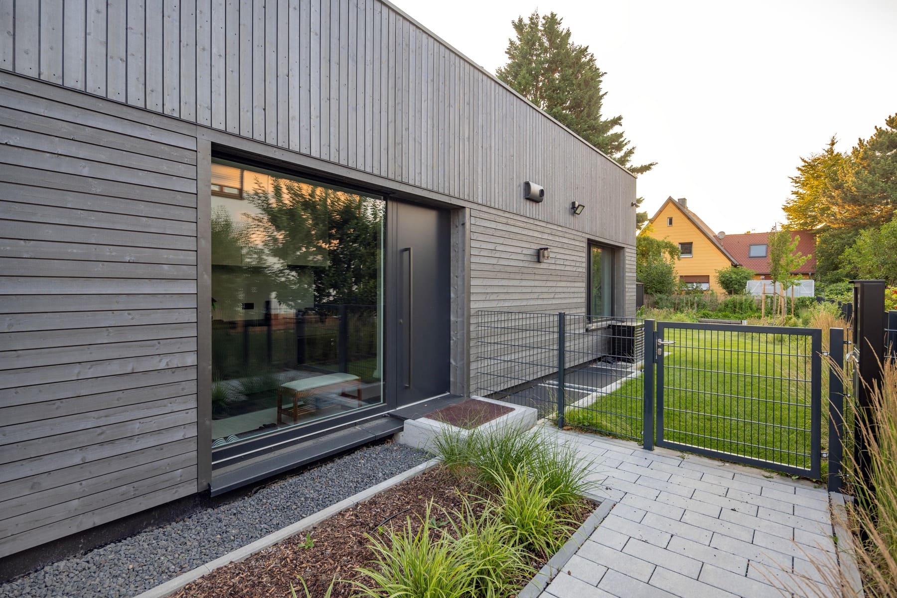 Moderne Holzfassade eines Hauses mit großen Fenstern und einem gepflegten Garten im Hintergrund.