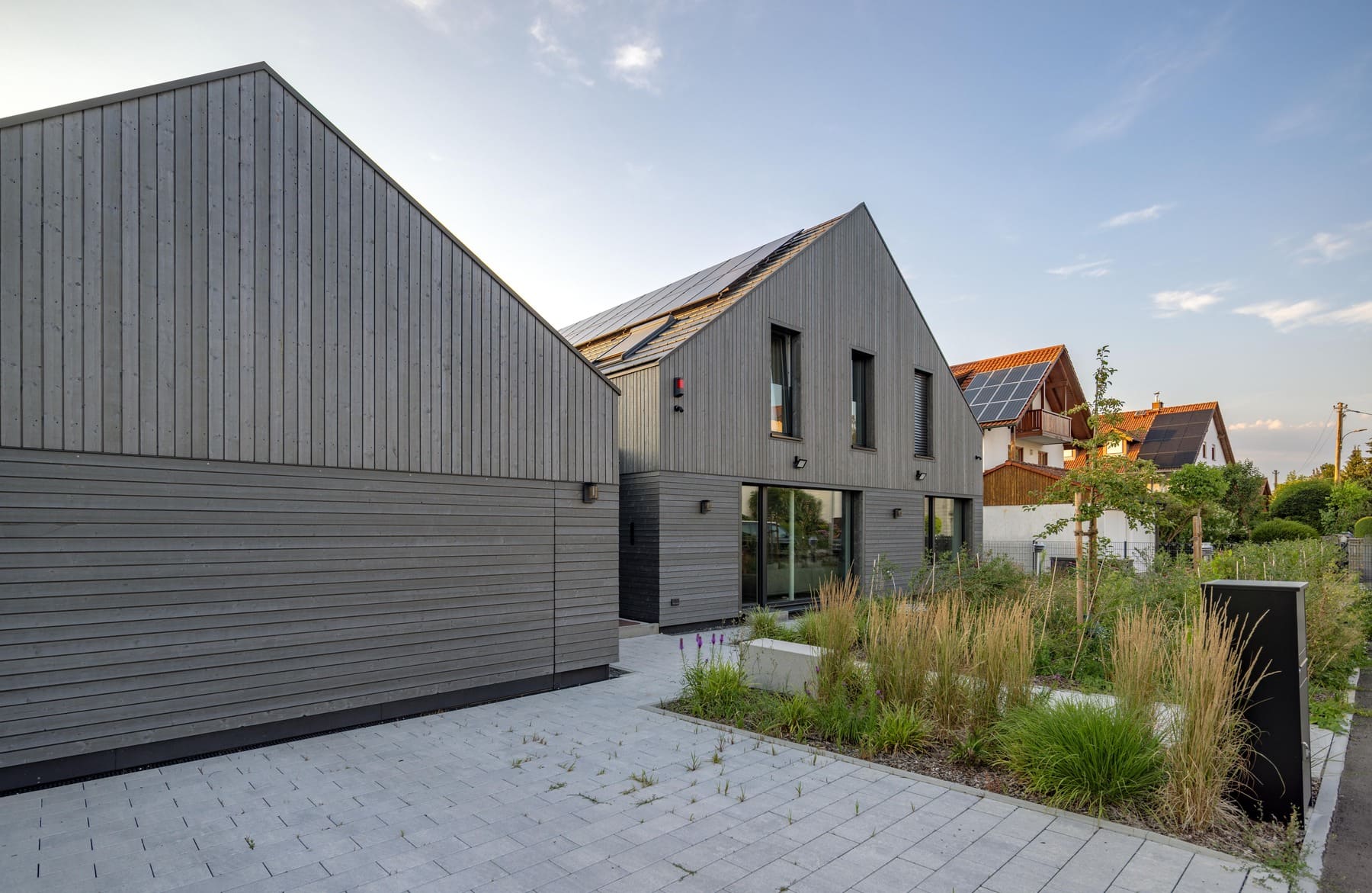 Modernes Haus mit Holzverkleidung und Garten, umgeben von grüner Landschaft und Nachbargebäuden.